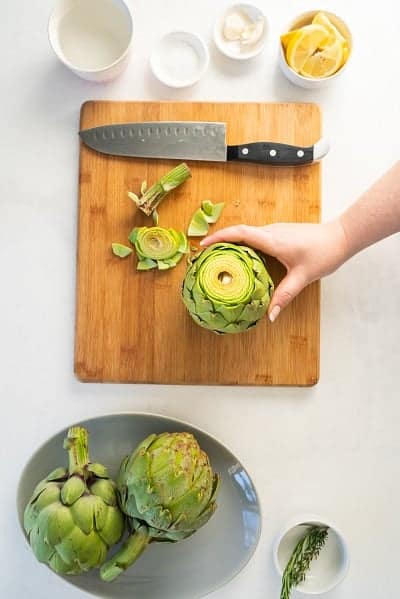 Top view photo of a hand holding an artichoke with the top cut off. It's on a wooden cutting board with a chefs knife next to it.
