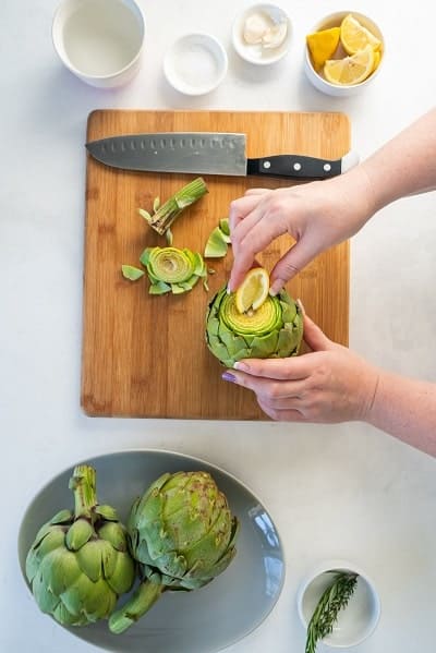 Top view photo of a hand squeezing lemon juice on the cut-side of the artichoke to prevent browning.