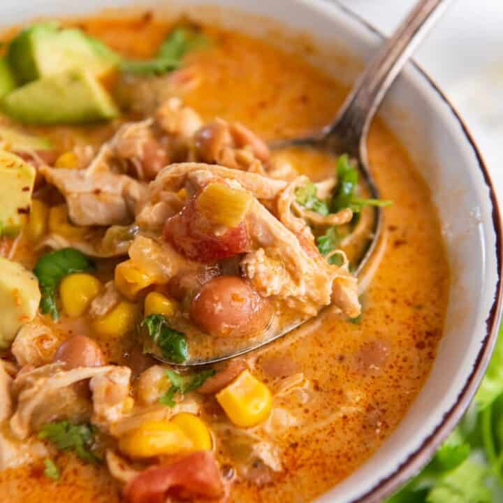 closeup shot of a white enameled bowl filled with brothy chicken chili with beans, corn, tomatoes, and cilantro. A spoon is lifting up a bite of the chili.