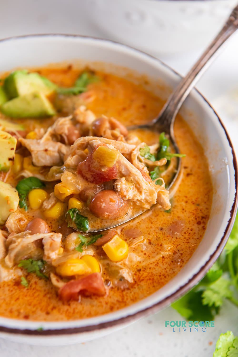 closeup shot of a white enameled bowl filled with brothy chicken chili with beans, corn, tomatoes, and cilantro. A spoon is lifting up a bite of the chili.