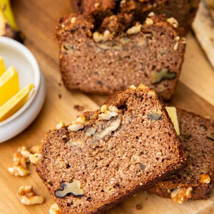 Slices of banana bread with walnuts on a wooden cutting board. a small bowl of butter pats is next to the bread.