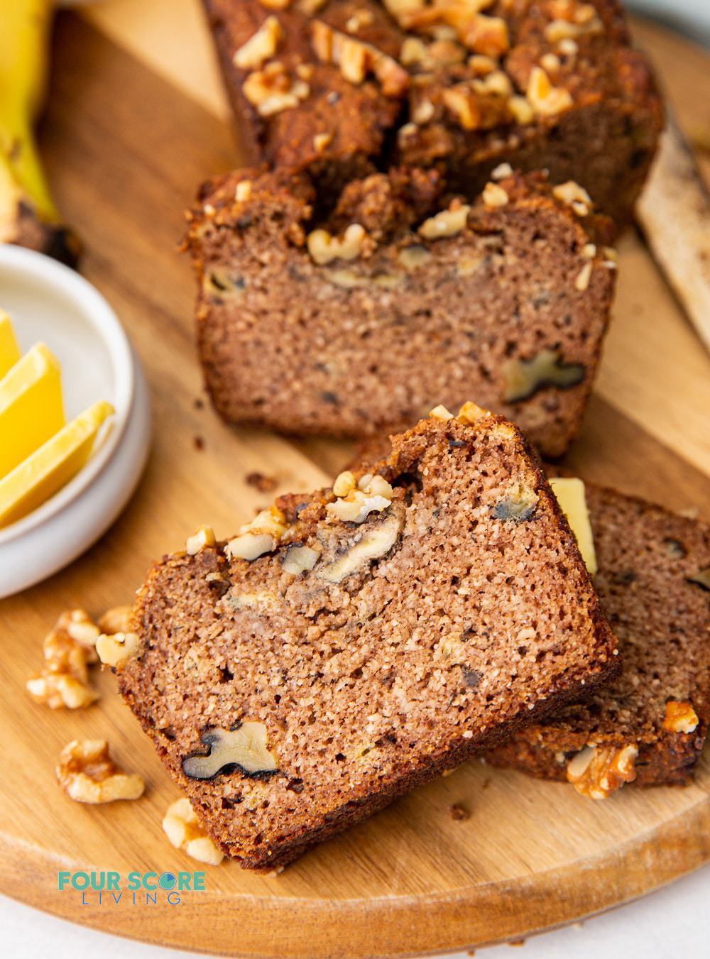 Slices of banana bread with walnuts on a wooden cutting board. a small bowl of butter pats is next to the bread.