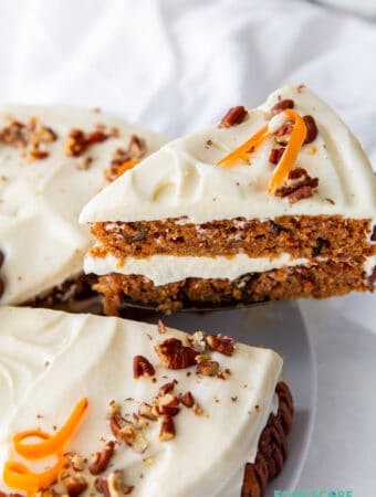 A wedge slice of two layer keto carrot cake being removed from the whole cake. Carrot cake is topped with frosting, chopped walnuts and carrot curls.