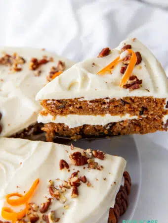 A wedge slice of two layer keto carrot cake being removed from the whole cake. Carrot cake is topped with frosting, chopped walnuts and carrot curls.