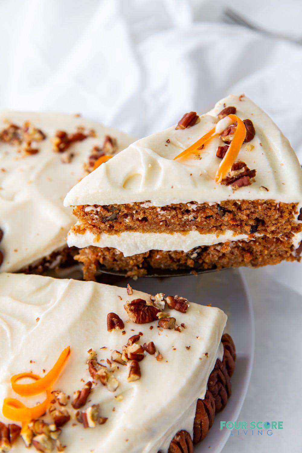 A wedge slice of two layer keto carrot cake being removed from the whole cake. Carrot cake is topped with frosting, chopped walnuts and carrot curls.