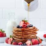 A tall stack of almond flour pancakes topped with fresh mixed berries and surrounded by fresh mixed berries. A hand is pouring syrup over the stack.