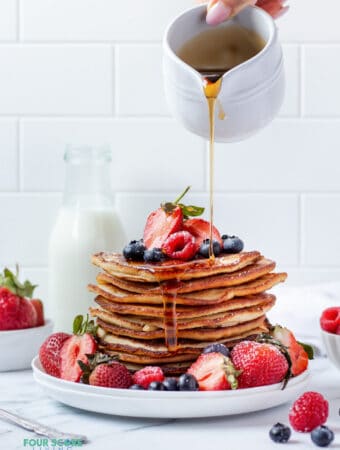 A tall stack of almond flour pancakes topped with fresh mixed berries and surrounded by fresh mixed berries. A hand is pouring syrup over the stack.