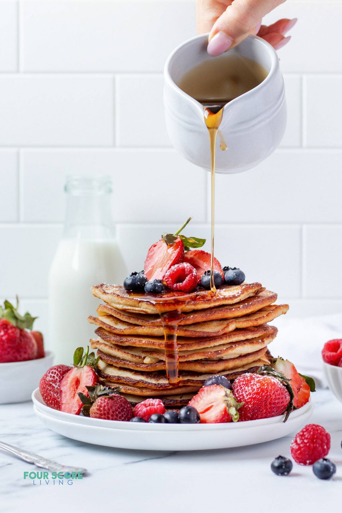 A tall stack of almond flour pancakes topped with fresh mixed berries and surrounded by fresh mixed berries. A hand is pouring syrup over the stack.