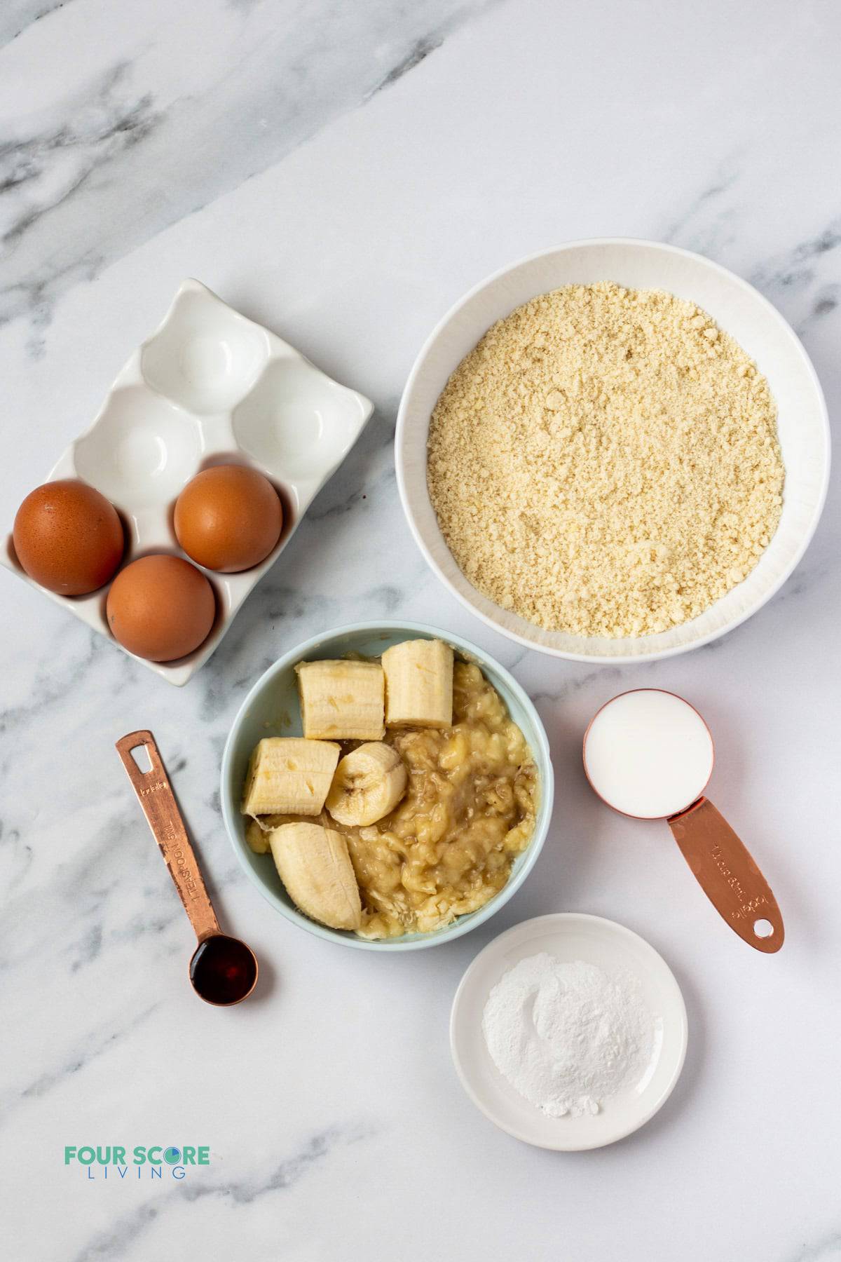 The ingredients needed to make almond flour banana pancakes, measured into separate bowls on a marble counter.