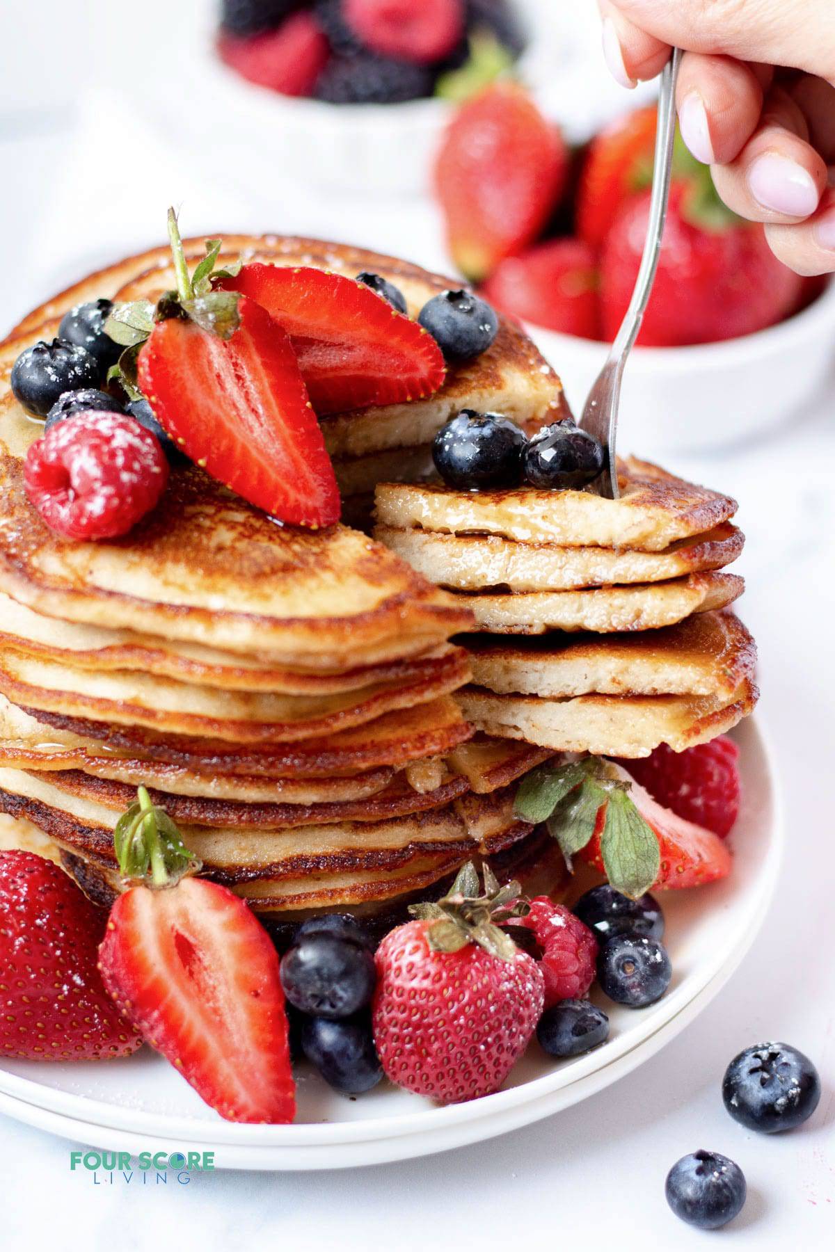 a tall stack of pancakes garnished with fresh berries. A fork is picking up a large bite of pancakes with blueberries on top.
