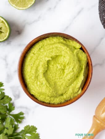 A bowl of green avocado crema on a marble counter, surrounded ty limes and cilantro