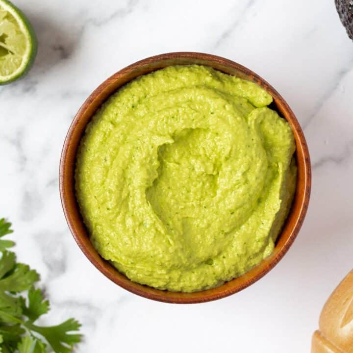 A bowl of green avocado crema on a marble counter, surrounded ty limes and cilantro