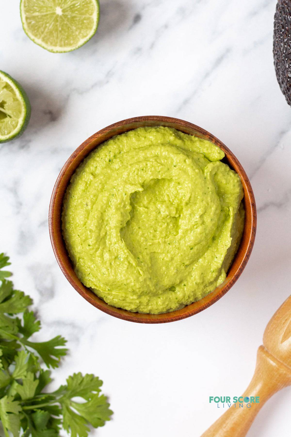 A bowl of green avocado crema on a marble counter, surrounded ty limes and cilantro