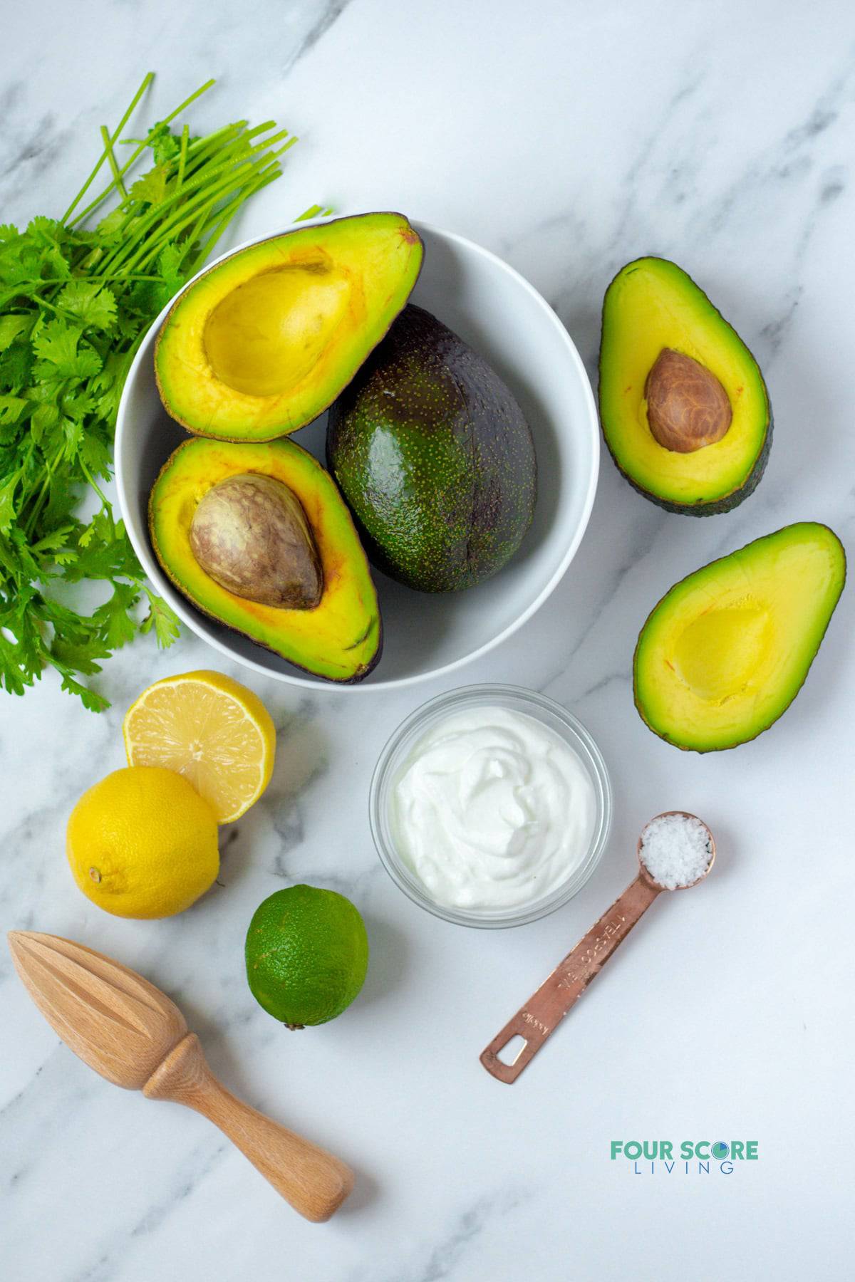 A bowl of halved avocados next to a small bowl of sour cream, a teaspoon of salt, a halved lemon, a whole lime, and a bunch of fresh cilantro