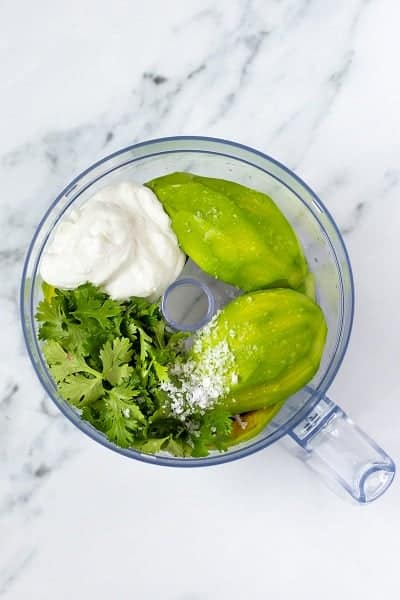 Top view photo of a blender with avocado flesh, cilantro, sour cream, salt, and lemon and lime juice, ready to be blended until smooth.