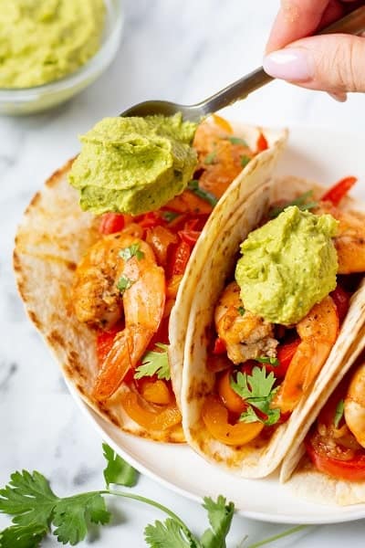 Closeup photo of a spoonful of avocado crema, being dalloped onto a couple shrimp tacos.