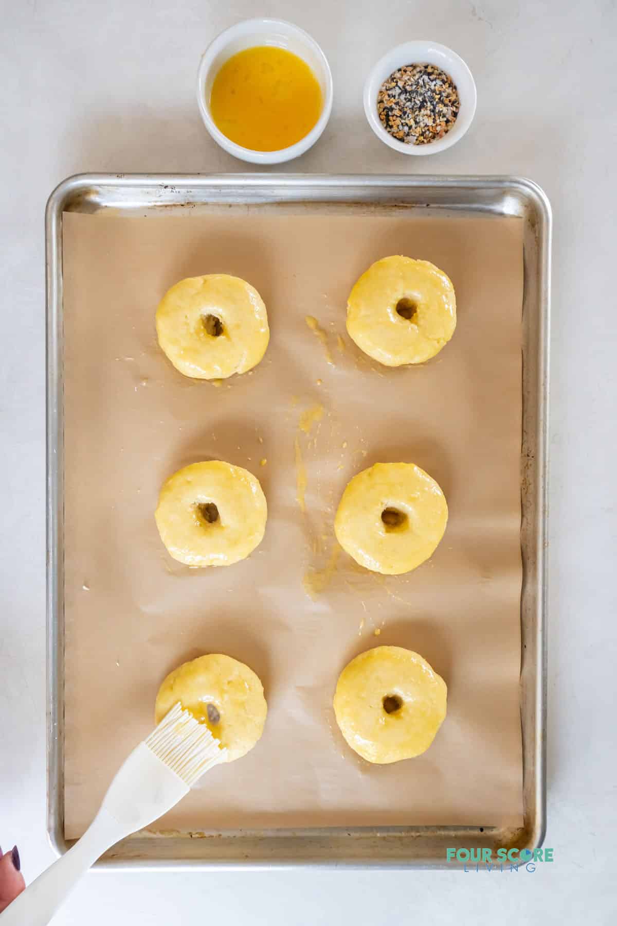 6 uncooked keto bagels on a tray. A silicone brush adds egg wash to them. A bowl of egg wash and a bowl of bagel seasoning are next to the pan.
