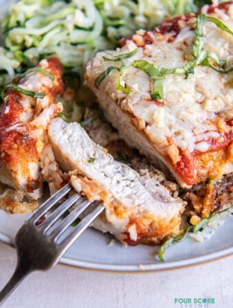 a dinner plate of keto chicken parmesan and zucchini noodles. The chicken is sliced and a bite is held with a fork.