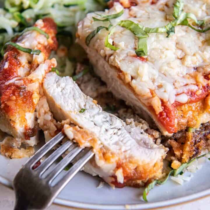 a dinner plate of keto chicken parmesan and zucchini noodles. The chicken is sliced and a bite is held with a fork.