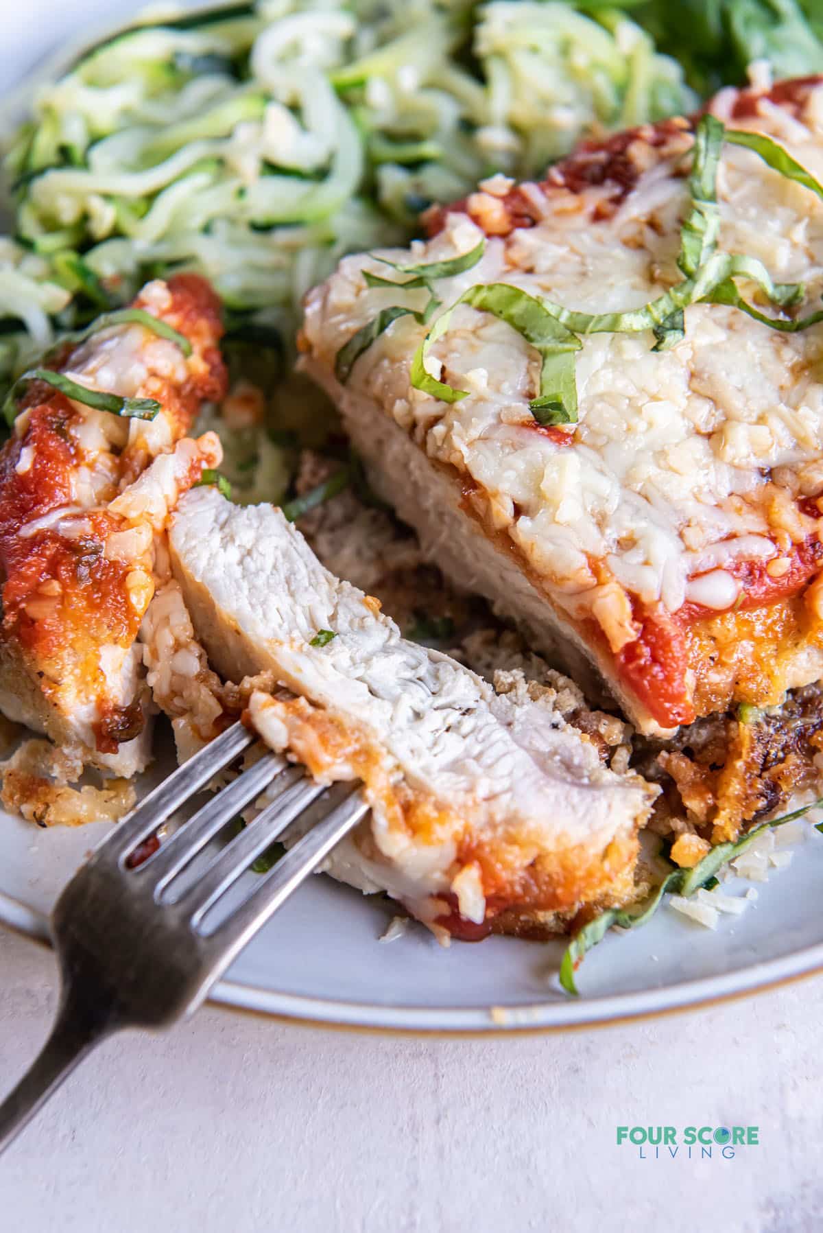 a dinner plate of keto chicken parmesan and zucchini noodles. The chicken is sliced and a bite is held with a fork.