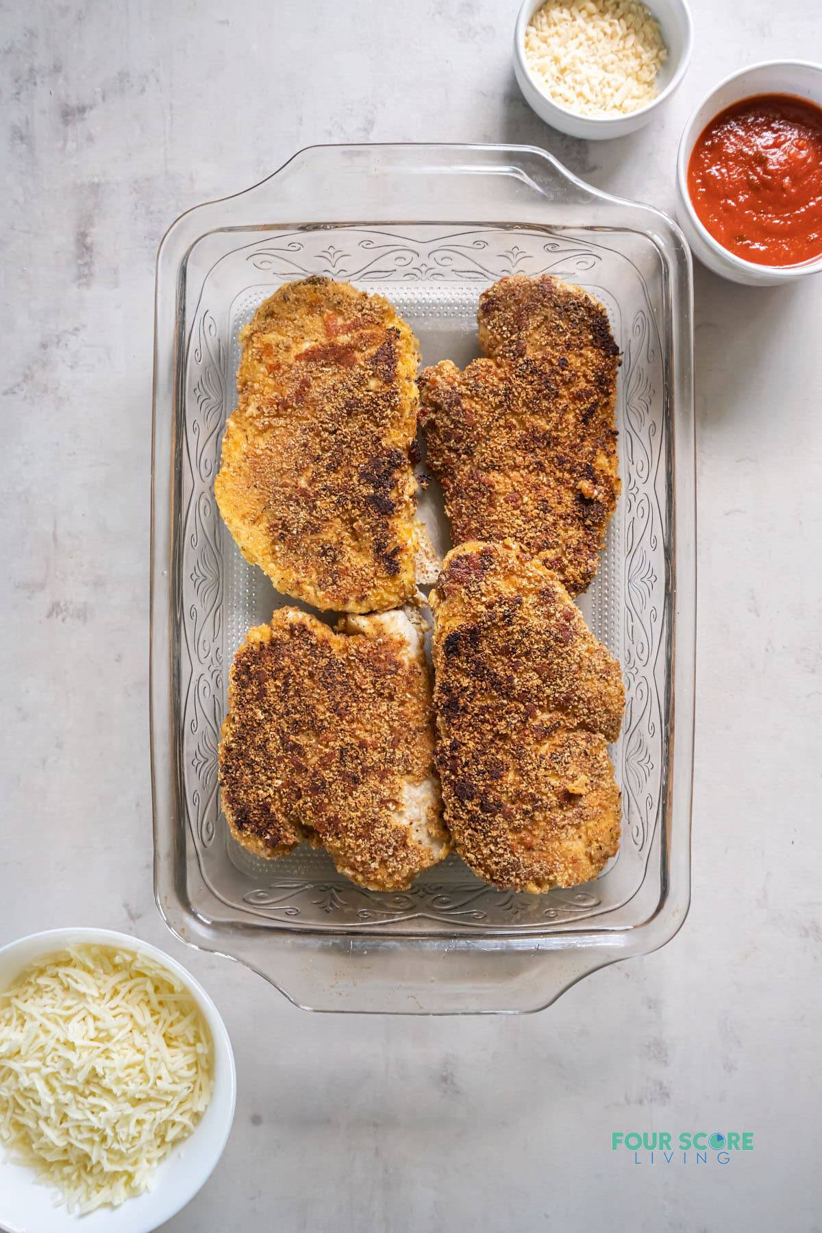 baked keto chicken parmesan cutlets in a glass baking dish. Bowls of cheese and sauce are nearby.