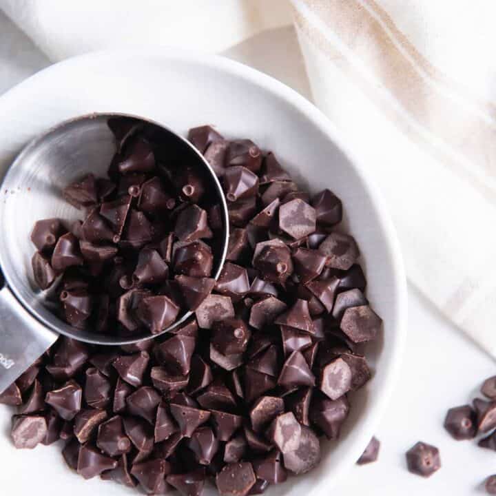 a bowl of homemade keto chocolate chips scooped by a small silver measuring cup, viewed from above