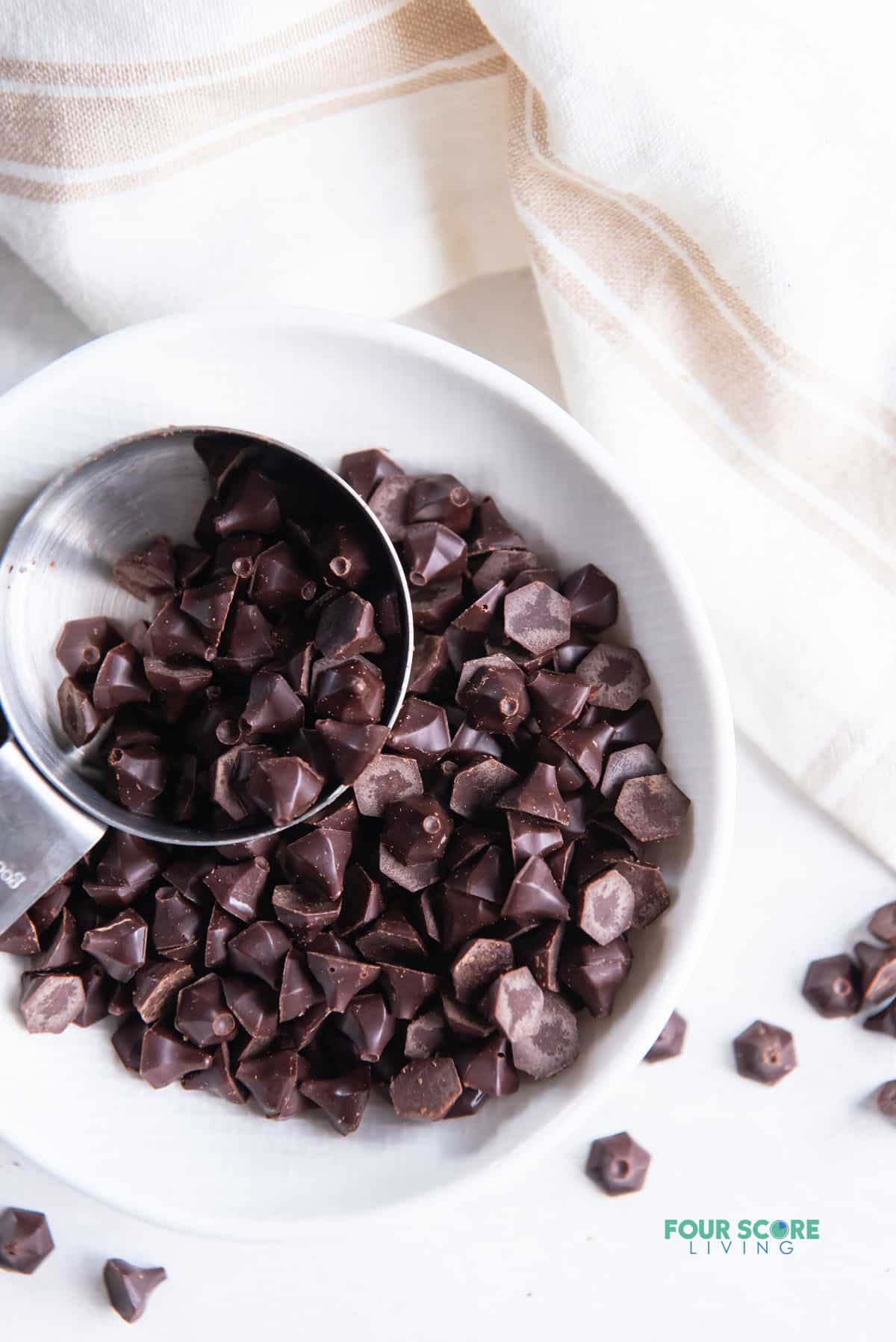 a bowl of homemade keto chocolate chips scooped by a small silver measuring cup, viewed from above