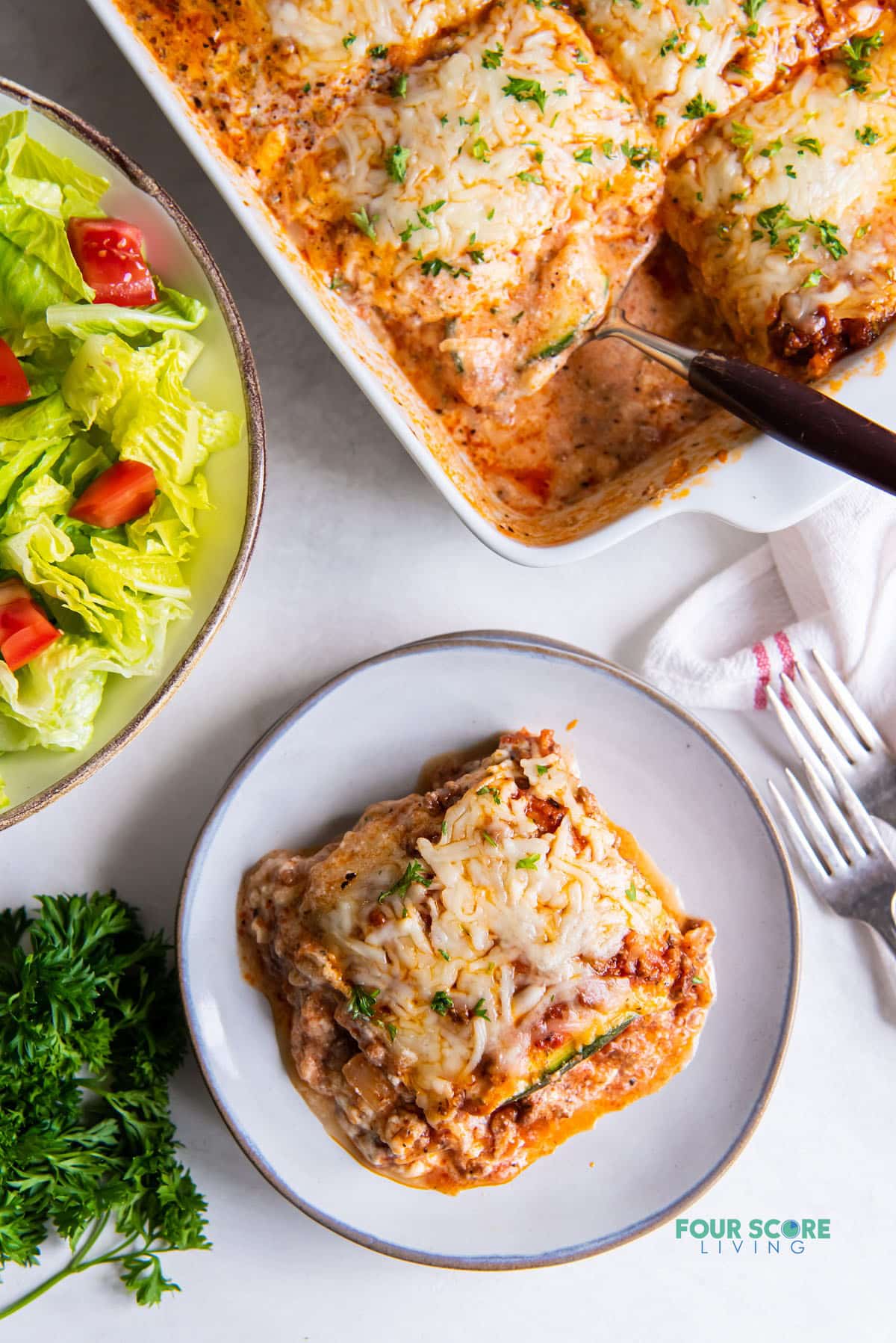 a serving of lasagna on a round white plate, next to the pan of lasagna and a side salad of lettuce and tomatoes