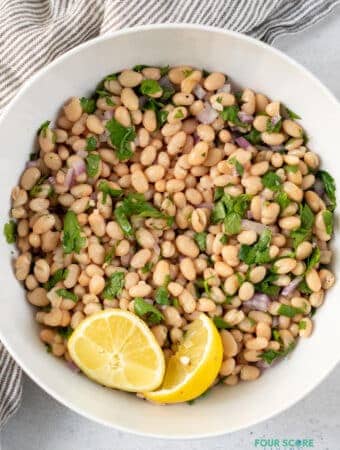 top view photo of white bean recipe in a white bowl with two lemon wedges, and a stripped kitchen towel above the bowl