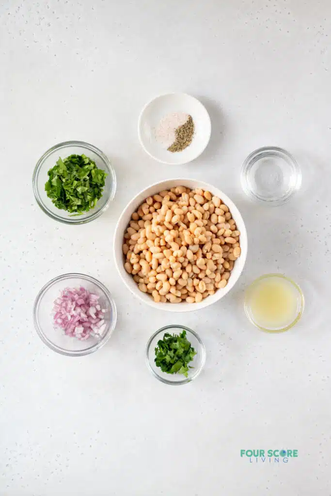 top view photo of ingredients for white bean recipe in separate bowls, including white beans,  red onion, cilantro, parsley, lemon juice, white vinegar, and salt and pepper