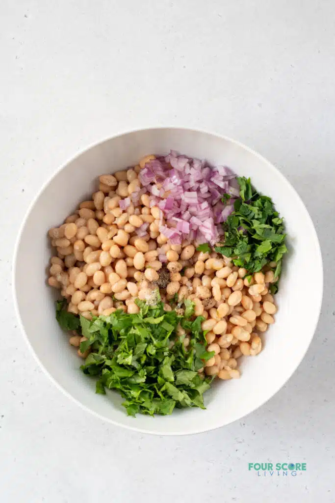 top view photo of ingredients to make white beans, all together in a large white bowl