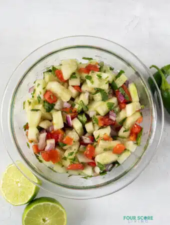 Top view photo of cucumber salsa in a clear class bowl with sliced limes and jalapenos around the bowl.