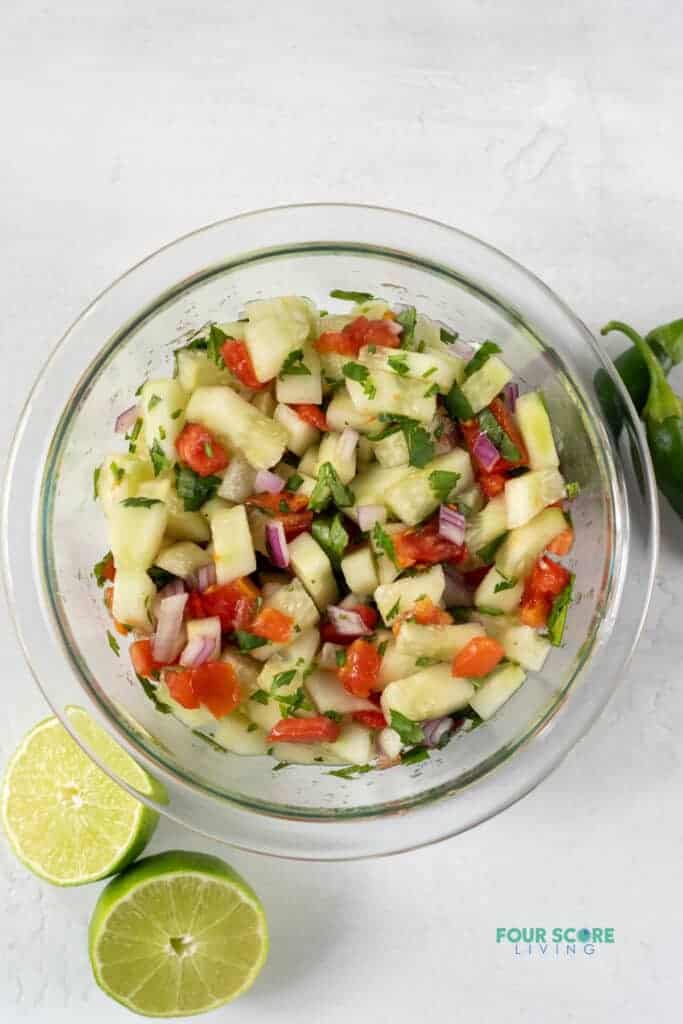 Top view photo of cucumber salsa in a clear class bowl with sliced limes and jalapenos around the bowl. 