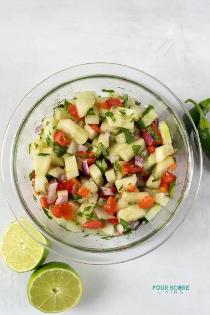 Top view photo of cucumber salsa in a clear class bowl with sliced limes and jalapenos around the bowl. 