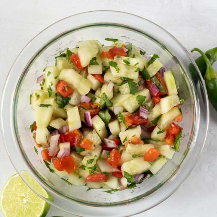 Top view photo of cucumber salsa in a clear class bowl with sliced limes and jalapenos around the bowl.