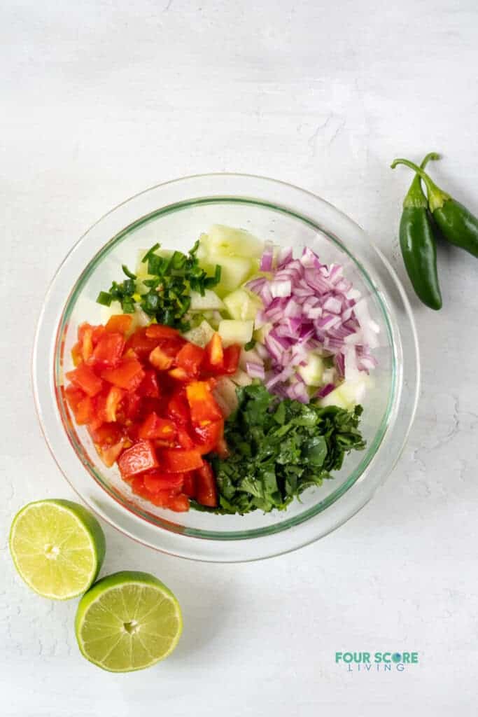 Top view photo of ingredients to make Cucumber Salsa in a clear glass bowl, ready to be mixed together. There are sliced limes and jalapenos by the bowl.