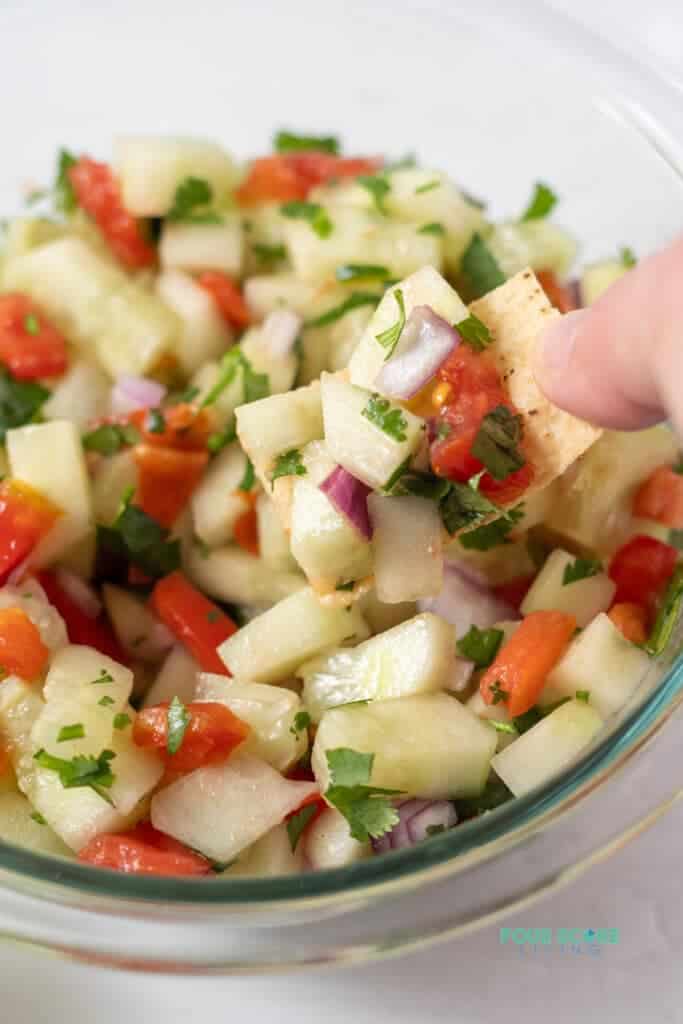 Closeup photo of a hand dipping a tortilla chip into a clear glass bowl filled with cucumber salsa.