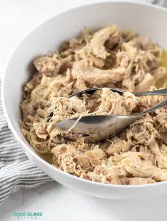 Photo of Instant Pot Shredded Chicken in a white bowl with silver tongs, ready to be served.