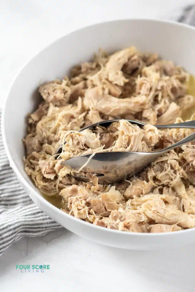 Photo of Instant Pot Shredded Chicken in a white bowl with silver tongs, ready to be served. 
