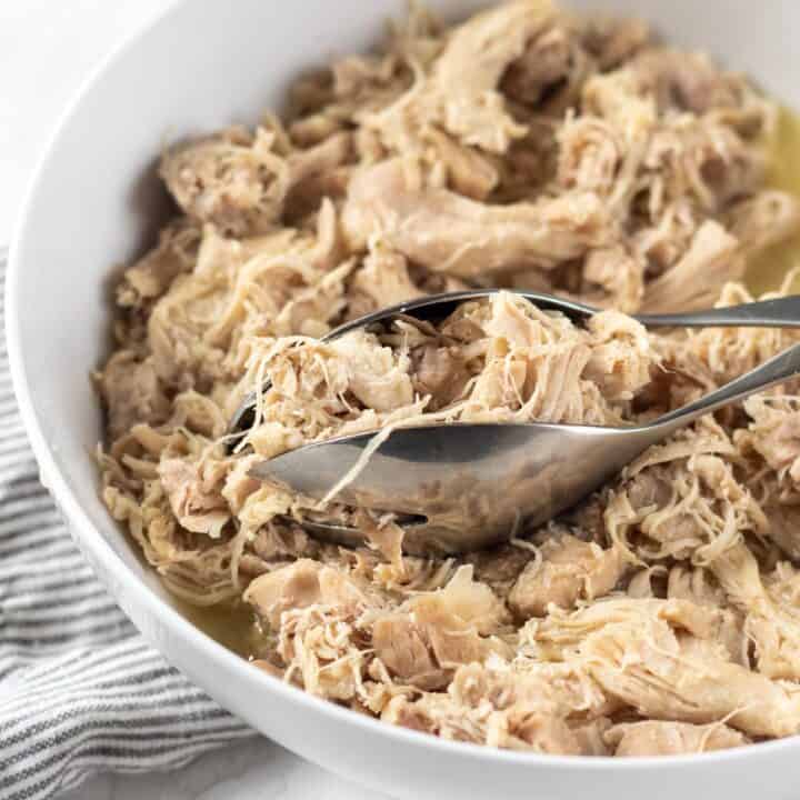 Photo of Instant Pot Shredded Chicken in a white bowl with silver tongs, ready to be served.