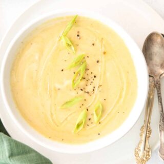 Top view photo of Magic Leek Soup in a white bowl, on a white plate. There are two silver spoons on the plate and a green kitchen towel by the bowl.