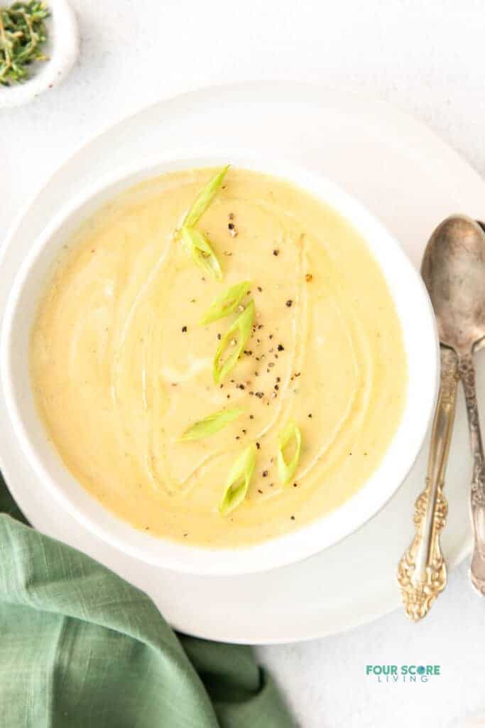 Top view photo of Magic Leek Soup in a white bowl, on a white plate. There are two silver spoons on the plate and a green kitchen towel by the bowl. 