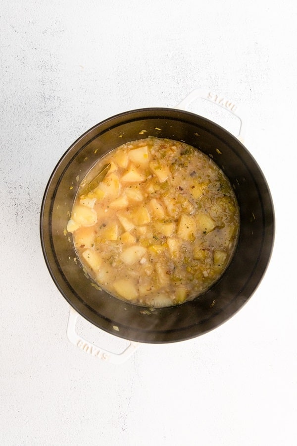 Top view photo of a large stockpot with sliced leeks, garlic, potatoes, broth, thyme, and bay leaf, simmering.