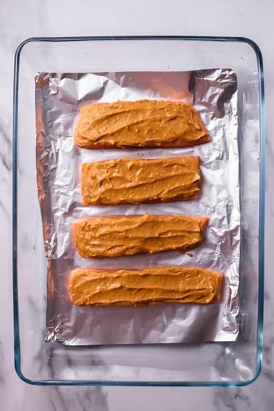 Top view photo of 4 salmon fillets in a clear baking dish lined with tin foil. The salmon has a peanut butter glaze spread on top.