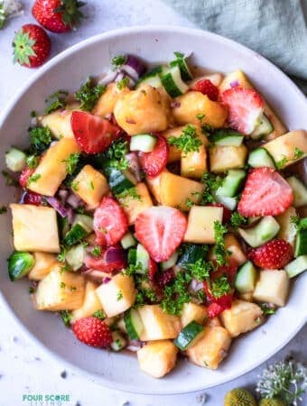 top view photo of pineapple salad in a white bowl, with strawberries and a kitchen towel above the bowl and flowers below it