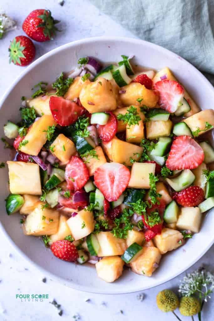 top view photo of pineapple salad in a white bowl, with strawberries and a kitchen towel above the bowl and flowers below it