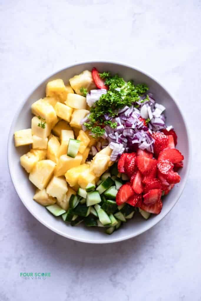 top view photo of ingredients to make pineapple salad in a white bowl, ready to be mixed, including pineapple, cucumber, strawberries, red onion, and cilantro.