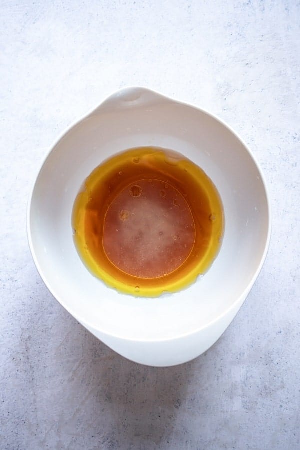 top view photo of a white bowl with ingredients to make the dressing for pineapple salad, including olive oil, honey, vinegar, lime juice, and salt
