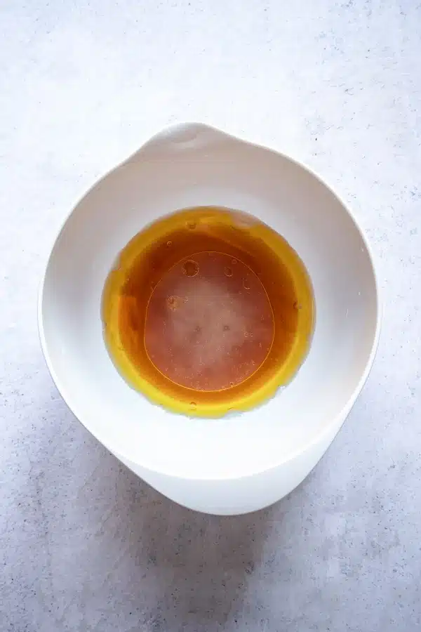 top view photo of a white bowl with ingredients to make the dressing for pineapple salad, including olive oil, honey, vinegar, lime juice, and salt