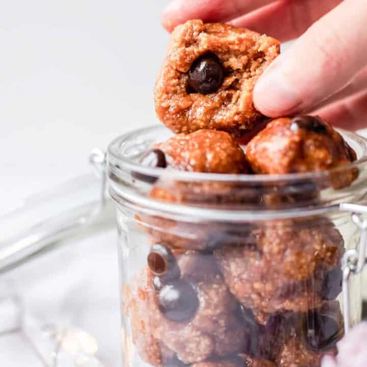 closeup photo of a hand grabbing a protein cookie dough ball with a bite taken out of it, in a glass jar.